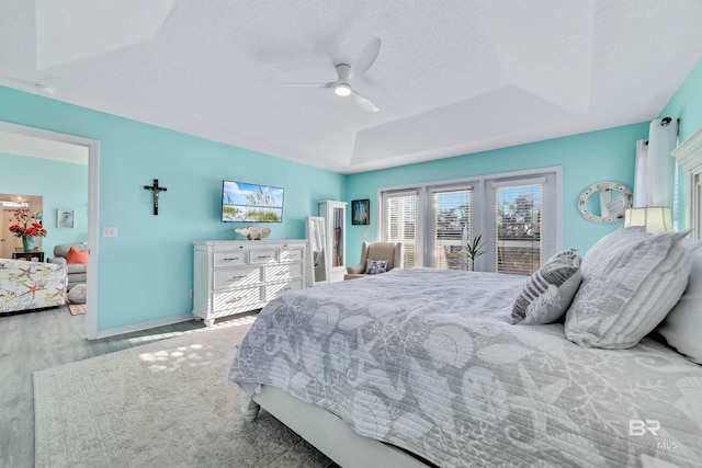 bedroom with a textured ceiling, ceiling fan, wood finished floors, baseboards, and a tray ceiling