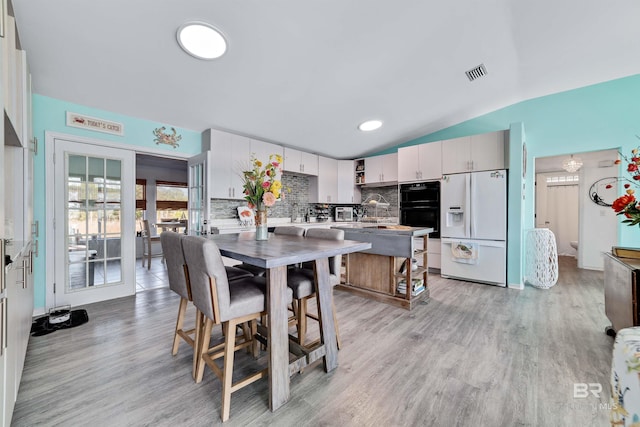 kitchen featuring white refrigerator with ice dispenser, visible vents, white cabinets, vaulted ceiling, and decorative backsplash