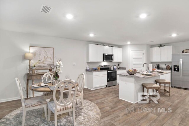 kitchen with a kitchen island with sink, white cabinetry, light stone counters, wood-type flooring, and stainless steel appliances