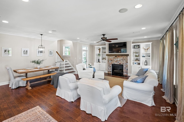 living area featuring built in shelves, a fireplace, crown molding, dark wood finished floors, and ceiling fan with notable chandelier