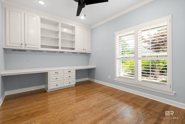 unfurnished office with built in desk, light wood finished floors, a ceiling fan, baseboards, and ornamental molding
