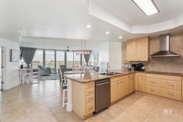 kitchen with a peninsula, a sink, wall chimney range hood, stainless steel dishwasher, and light brown cabinetry