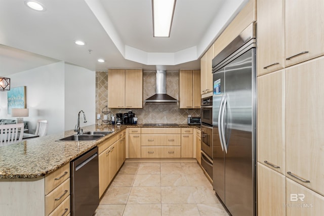 kitchen featuring a peninsula, appliances with stainless steel finishes, wall chimney exhaust hood, light stone countertops, and light brown cabinetry