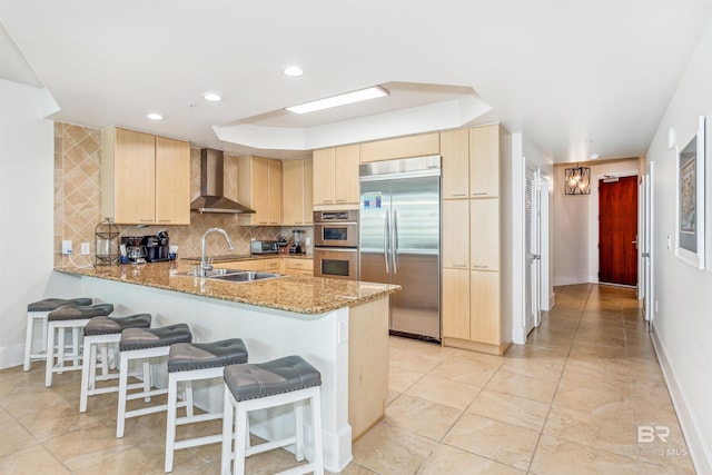 kitchen with stainless steel appliances, a peninsula, a sink, wall chimney range hood, and light brown cabinetry