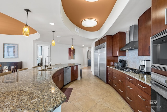 kitchen featuring sink, an island with sink, wall chimney range hood, stainless steel appliances, and decorative light fixtures