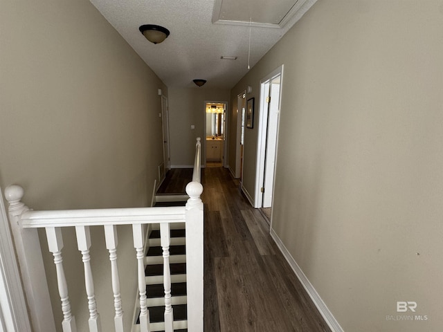 corridor with dark wood-style flooring, a textured ceiling, attic access, baseboards, and an upstairs landing