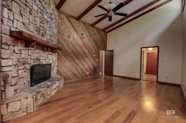 unfurnished living room featuring beam ceiling, high vaulted ceiling, light hardwood / wood-style floors, a stone fireplace, and wood walls