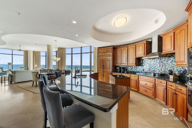 kitchen featuring decorative backsplash, wall chimney exhaust hood, a tray ceiling, an island with sink, and a kitchen bar