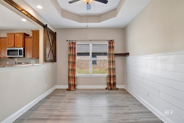 empty room with light hardwood / wood-style floors, a raised ceiling, a barn door, and ceiling fan