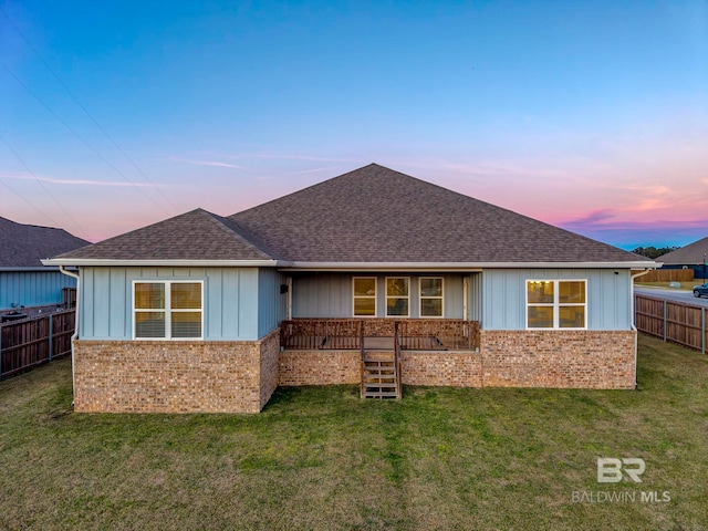 back of house at dusk with a yard, brick siding, a shingled roof, and fence