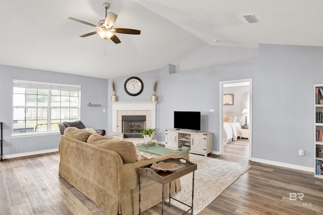 living area featuring a ceiling fan, a tiled fireplace, vaulted ceiling, wood finished floors, and baseboards