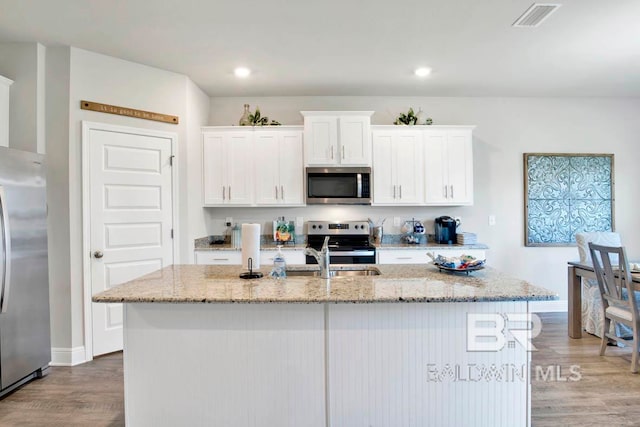 kitchen featuring stainless steel appliances, light stone countertops, an island with sink, white cabinets, and light hardwood / wood-style flooring