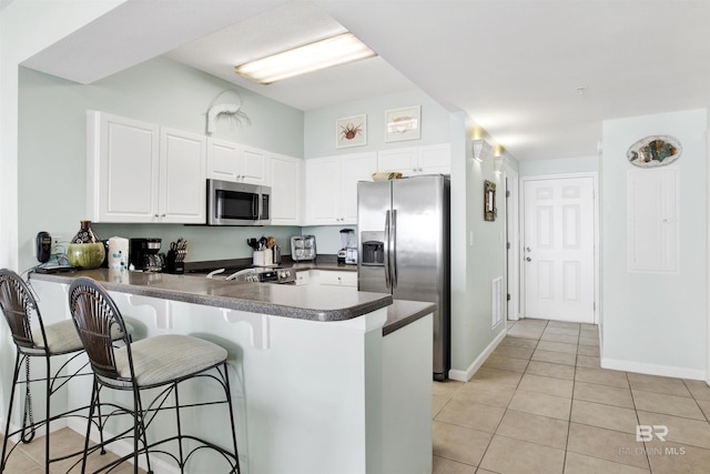 kitchen featuring light tile patterned flooring, stainless steel appliances, a peninsula, dark countertops, and a kitchen bar