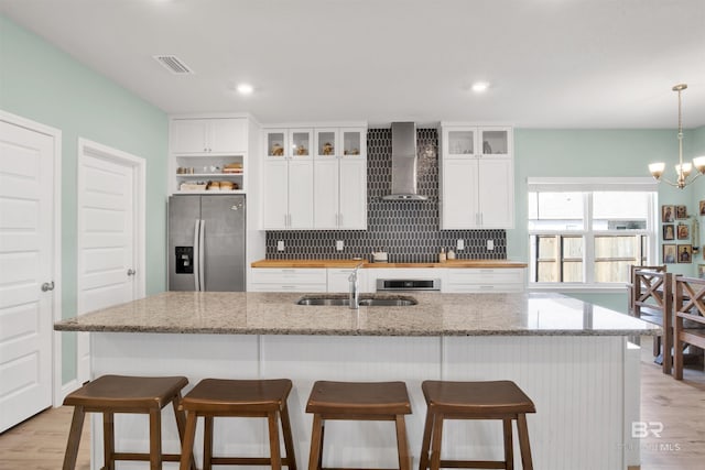 kitchen with sink, a kitchen island with sink, white cabinets, stainless steel fridge with ice dispenser, and wall chimney exhaust hood