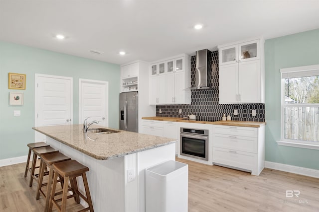 kitchen with an island with sink, sink, white cabinets, stainless steel appliances, and wall chimney exhaust hood