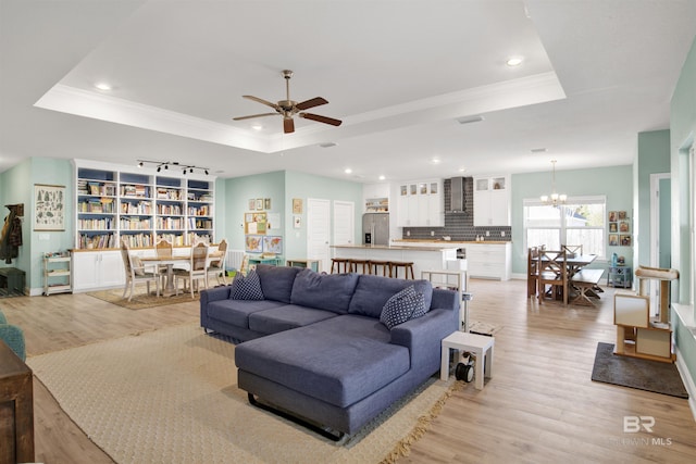 living room featuring crown molding, ceiling fan with notable chandelier, a raised ceiling, and light wood-type flooring