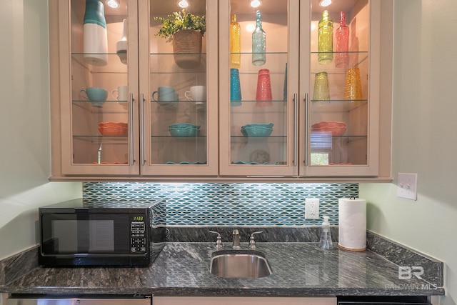 kitchen featuring a sink, dark stone countertops, backsplash, black microwave, and glass insert cabinets