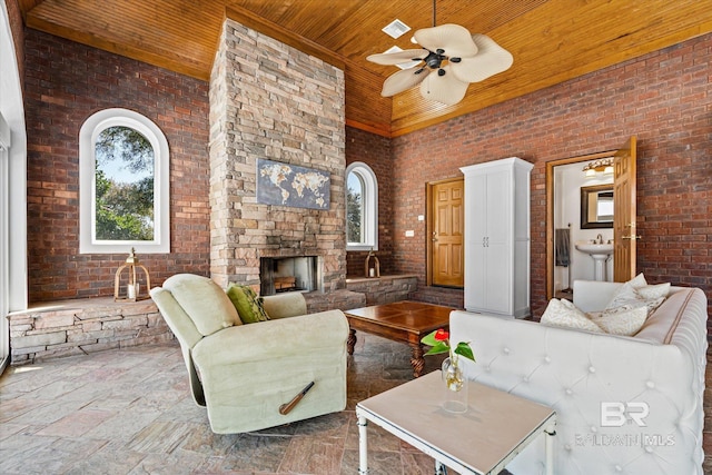 living area featuring a ceiling fan, an outdoor stone fireplace, wooden ceiling, brick wall, and a towering ceiling