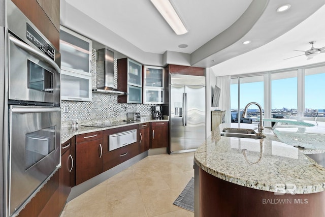 kitchen with wall chimney exhaust hood, stainless steel appliances, sink, ceiling fan, and a center island with sink