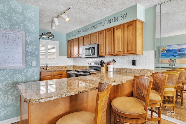 kitchen with a textured ceiling, appliances with stainless steel finishes, a peninsula, wallpapered walls, and light stone countertops