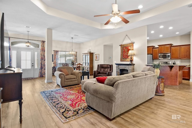 living room featuring a tray ceiling, light hardwood / wood-style flooring, a stone fireplace, and ceiling fan with notable chandelier