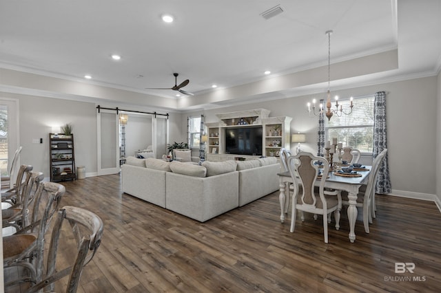 living room with ceiling fan with notable chandelier, a raised ceiling, crown molding, a barn door, and dark wood-type flooring