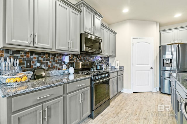 kitchen featuring gray cabinetry, stainless steel appliances, backsplash, dark stone counters, and light wood-type flooring