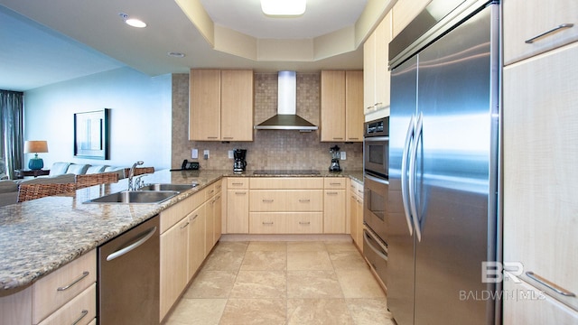 kitchen featuring light brown cabinets, appliances with stainless steel finishes, wall chimney exhaust hood, a sink, and a peninsula
