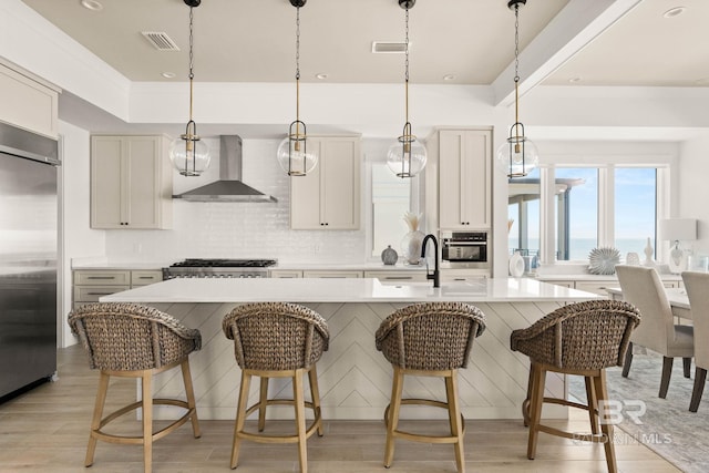 kitchen featuring wall chimney range hood, light hardwood / wood-style flooring, hanging light fixtures, a center island with sink, and stainless steel appliances