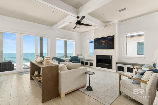 living room featuring a large fireplace, beamed ceiling, light wood-type flooring, a water view, and ceiling fan