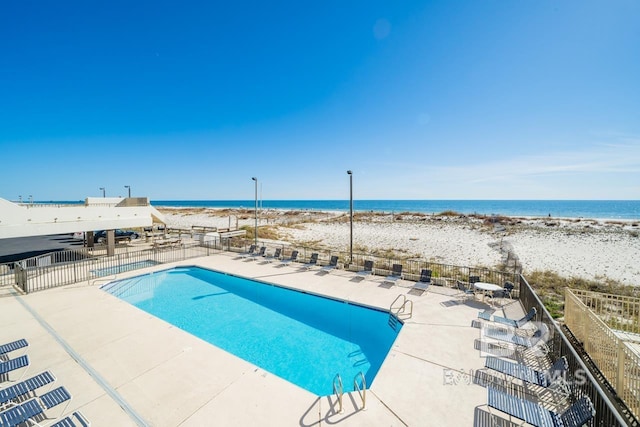 pool with a patio area, a water view, fence, and a beach view