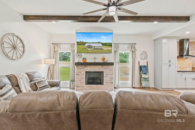 living room with ceiling fan, beam ceiling, a brick fireplace, and light hardwood / wood-style flooring