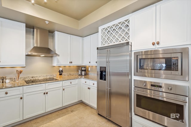 kitchen featuring wall chimney range hood, light stone countertops, built in appliances, and white cabinetry