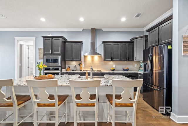 kitchen with wood finished floors, a kitchen island with sink, decorative backsplash, appliances with stainless steel finishes, and wall chimney range hood