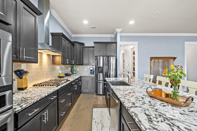 kitchen featuring light stone counters, a sink, appliances with stainless steel finishes, wall chimney exhaust hood, and backsplash