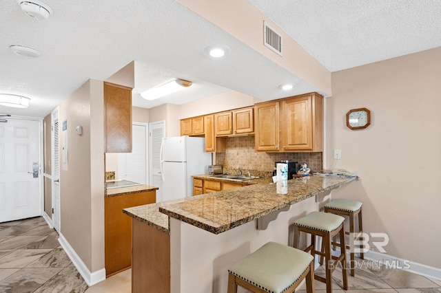 kitchen with white fridge, kitchen peninsula, light stone counters, sink, and backsplash