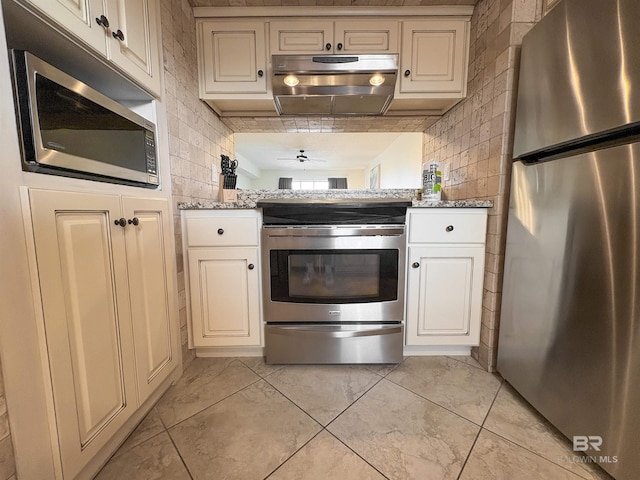 kitchen featuring light stone countertops, stainless steel appliances, decorative backsplash, cream cabinetry, and under cabinet range hood