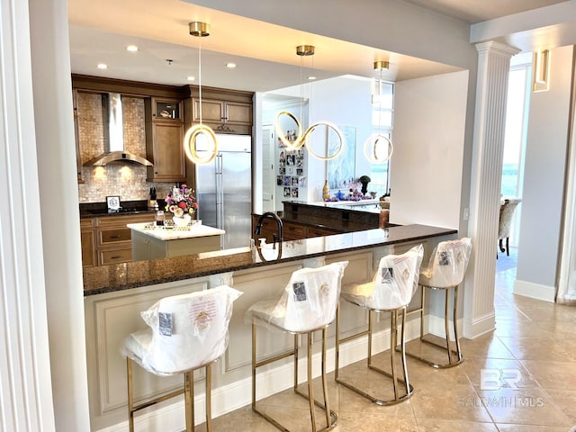 bar with dark stone countertops, stainless steel built in refrigerator, tasteful backsplash, wall chimney range hood, and pendant lighting