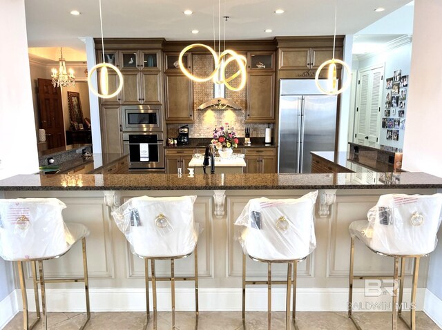 kitchen featuring built in appliances, decorative light fixtures, dark stone countertops, and backsplash