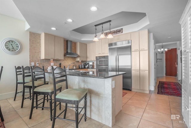 kitchen with a kitchen breakfast bar, a raised ceiling, wall chimney range hood, appliances with stainless steel finishes, and kitchen peninsula