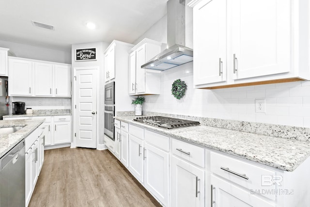 kitchen featuring white cabinetry, stainless steel appliances, light stone counters, wall chimney exhaust hood, and light wood-type flooring