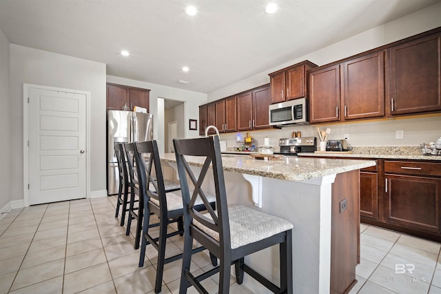 kitchen featuring appliances with stainless steel finishes, a kitchen breakfast bar, light tile patterned flooring, an island with sink, and light stone counters