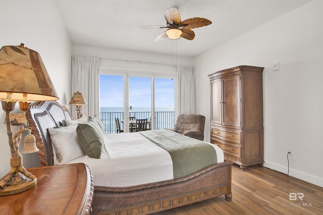 bedroom with a textured ceiling, access to outside, ceiling fan, a water view, and hardwood / wood-style floors