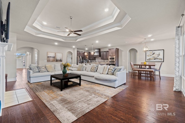 living room featuring ceiling fan with notable chandelier, a raised ceiling, ornamental molding, and dark wood-type flooring