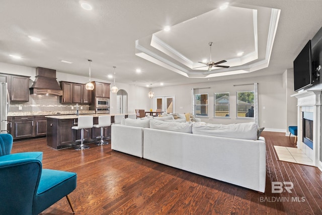 living room featuring dark wood-type flooring, crown molding, ceiling fan, a fireplace, and a tray ceiling