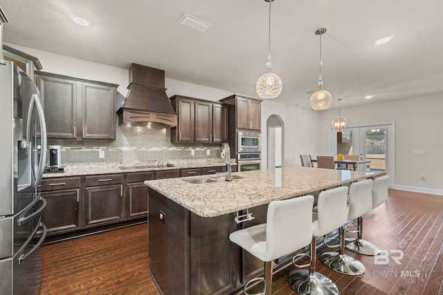 kitchen with appliances with stainless steel finishes, custom exhaust hood, sink, decorative light fixtures, and a center island with sink