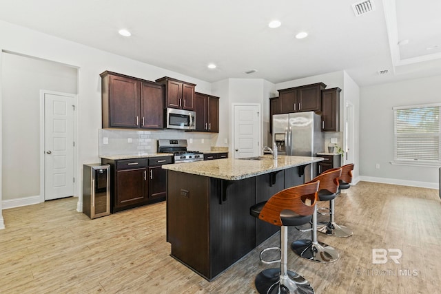 kitchen with appliances with stainless steel finishes, light stone countertops, light hardwood / wood-style floors, wine cooler, and a kitchen island with sink