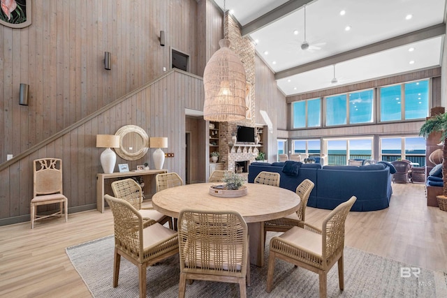 dining room featuring beam ceiling, light wood-type flooring, ceiling fan, a fireplace, and a high ceiling