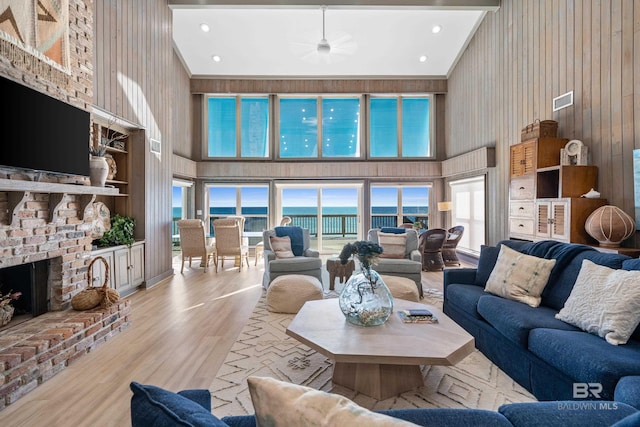 living room with high vaulted ceiling, wood walls, light hardwood / wood-style floors, a water view, and a brick fireplace
