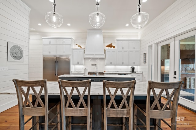 kitchen featuring a center island with sink, custom range hood, hanging light fixtures, a sink, and built in fridge